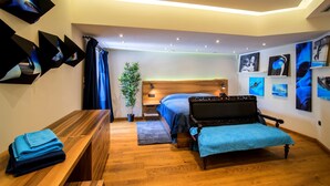 A modern bedroom with a wooden floor, a bed with a blue bedspread, a bench, a small table with towels, and wall art.
