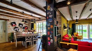 A rustic interior with exposed wooden beams, a stone wall, a dining area with a wooden table and chairs, a red sofa, and a bookshelf.