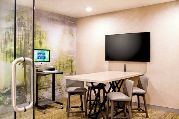 A conference room with a large table, four chairs, a mounted TV, a computer on a stand, and a glass partition with a view of trees.