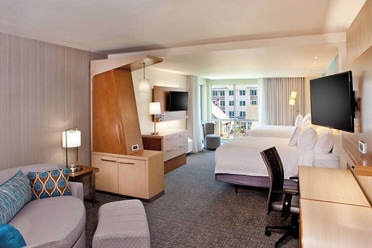 Quarto de hotel com uma cama, uma escrivaninha, uma cadeira, uma televisão e uma janela com vista para edifícios.