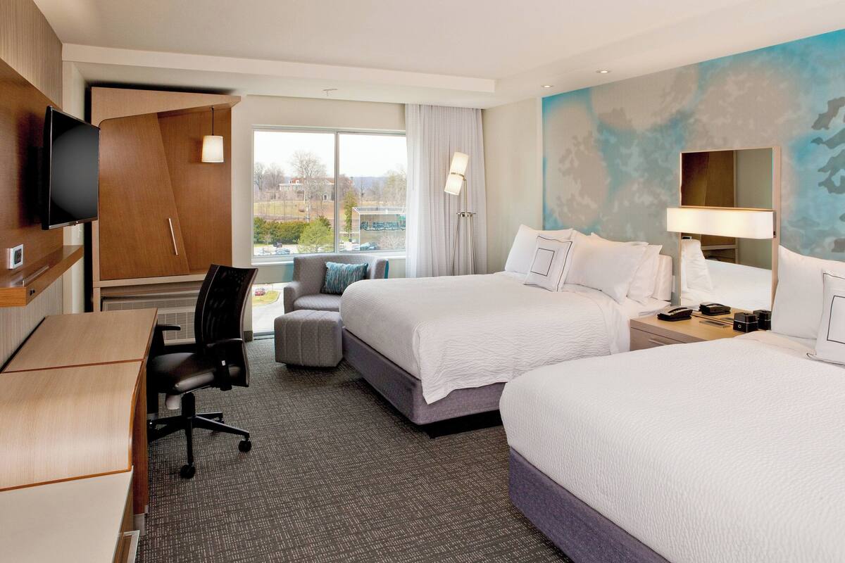 Quarto de hotel com duas camas, uma escrivaninha, uma cadeira, uma televisão e uma janela com vista para edifícios.