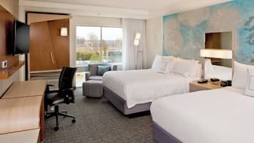 A hotel room with two beds, a desk, a chair, a TV, and a window with a view of buildings.