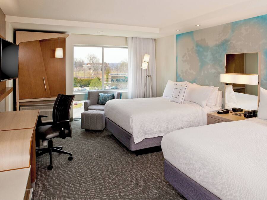 A hotel room with two beds, a desk, a chair, a TV, and a window with a view of buildings.