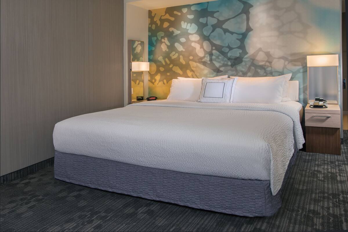 A hotel room with a large bed, a nightstand with a lamp, a wall with a textured design, and a carpeted floor.