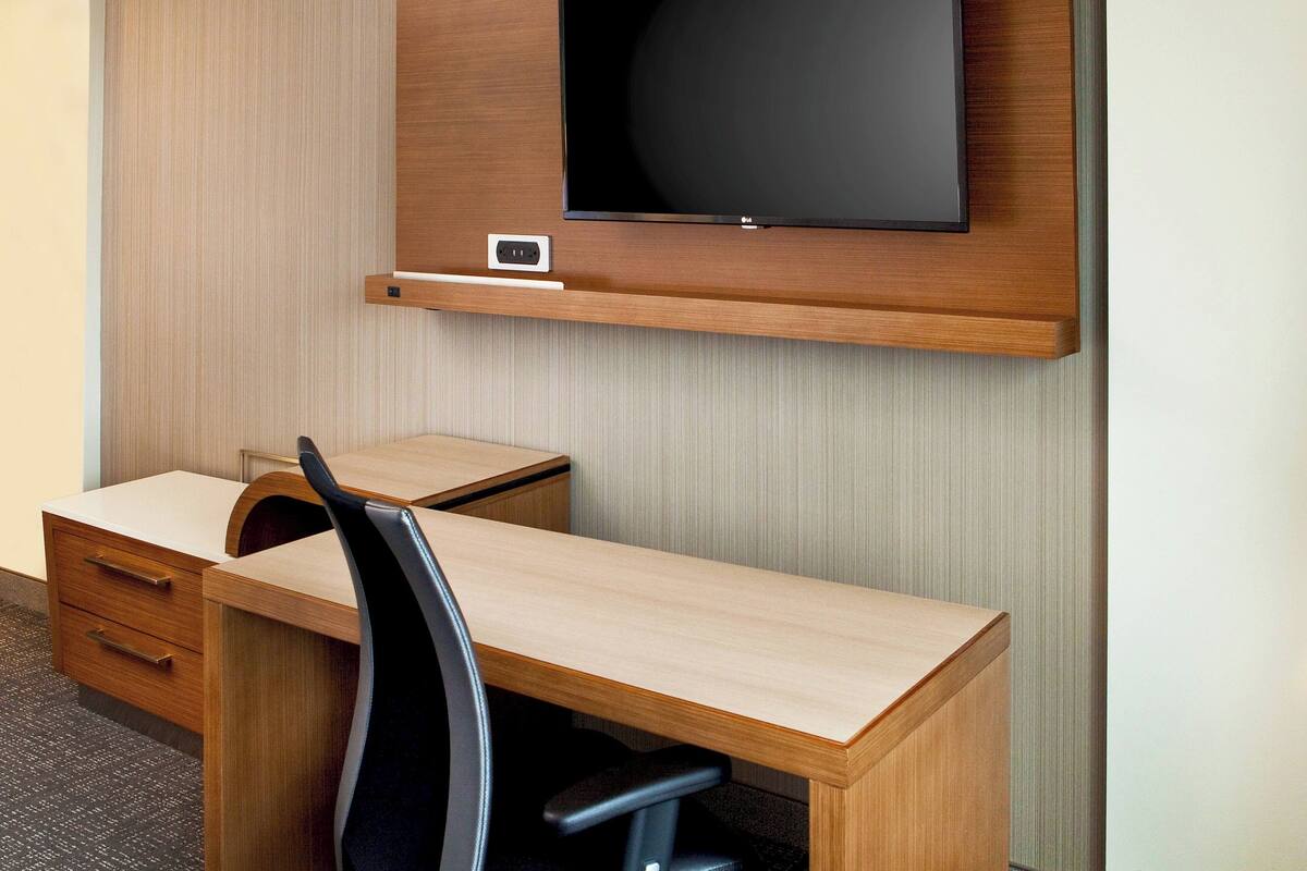 Quarto de hotel com uma escrivaninha de madeira, uma cadeira de escritório preta, uma televisão de tela plana fixada na parede e uma pequena prateleira abaixo da TV.