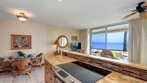 A modern kitchen with a large island, a dining area with wicker chairs, and a view of the ocean. - Pikake #a6 2 Bedroom Condo by RedAwning (Lahaina)