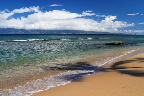 A beach with clear water, a sandy shore, and a distant mountain range under a blue sky with clouds.