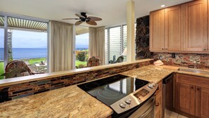 A kitchen with a large island, a view of the ocean, wooden cabinets, and a stone accent wall. - Pikake #a6 2 Bedroom Condo by RedAwning (Lahaina)