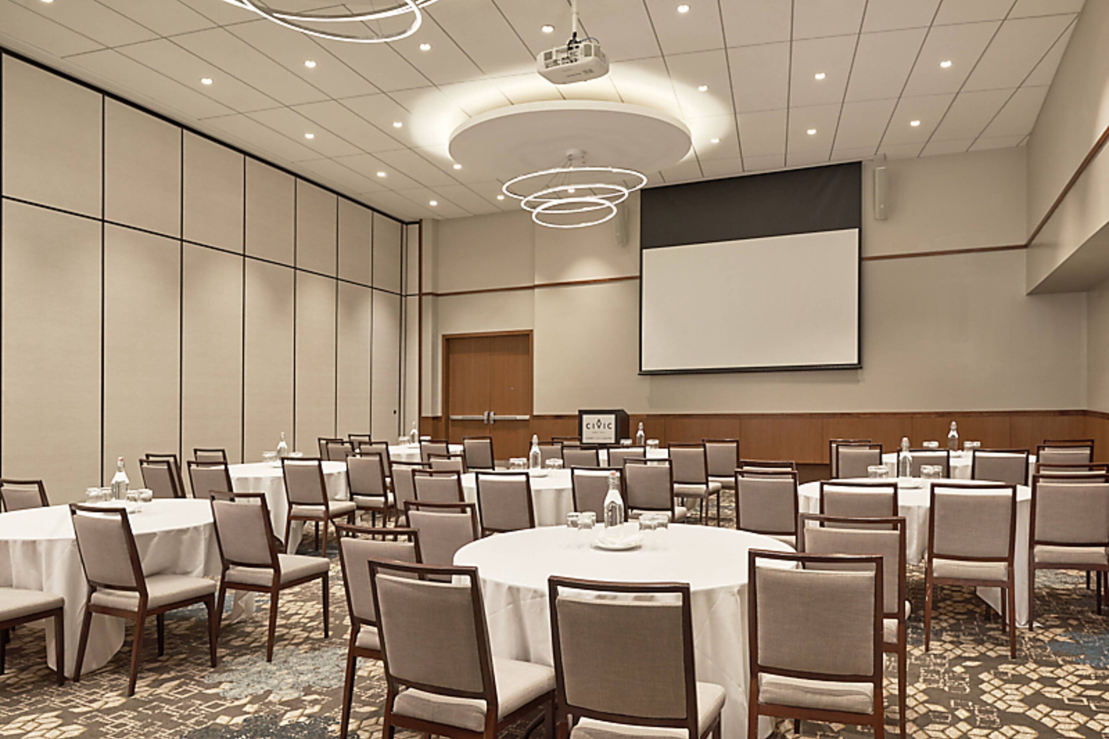 A conference room with multiple round tables, chairs, a projector screen, and a large whiteboard.