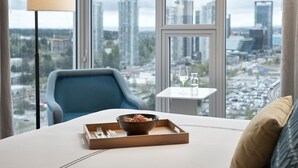 A hotel room with a large window offering a city view, a dining table with a tray of food, a blue armchair, and a floor lamp. - Civic Hotel Autograph Collection (Surrey)