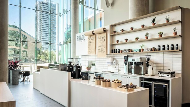 A modern cafe with a large window, a counter with coffee machines, shelves with potted plants, and a sign reading "PRADO CAFE". - Civic Hotel Autograph Collection (Surrey)