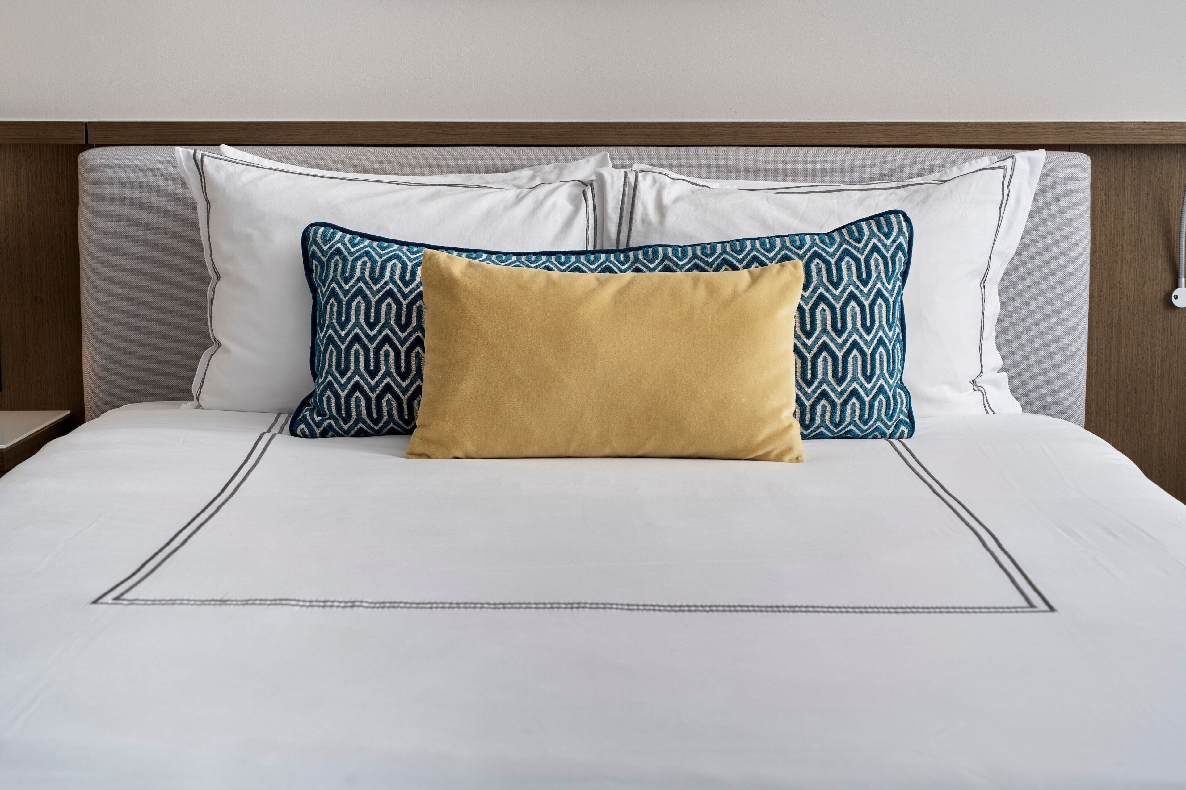 Ein ordentlich bezogenes Bett mit weißen Leinen und einem goldenen Kissen in der Mitte, begleitet von zwei Kissen mit Muster.