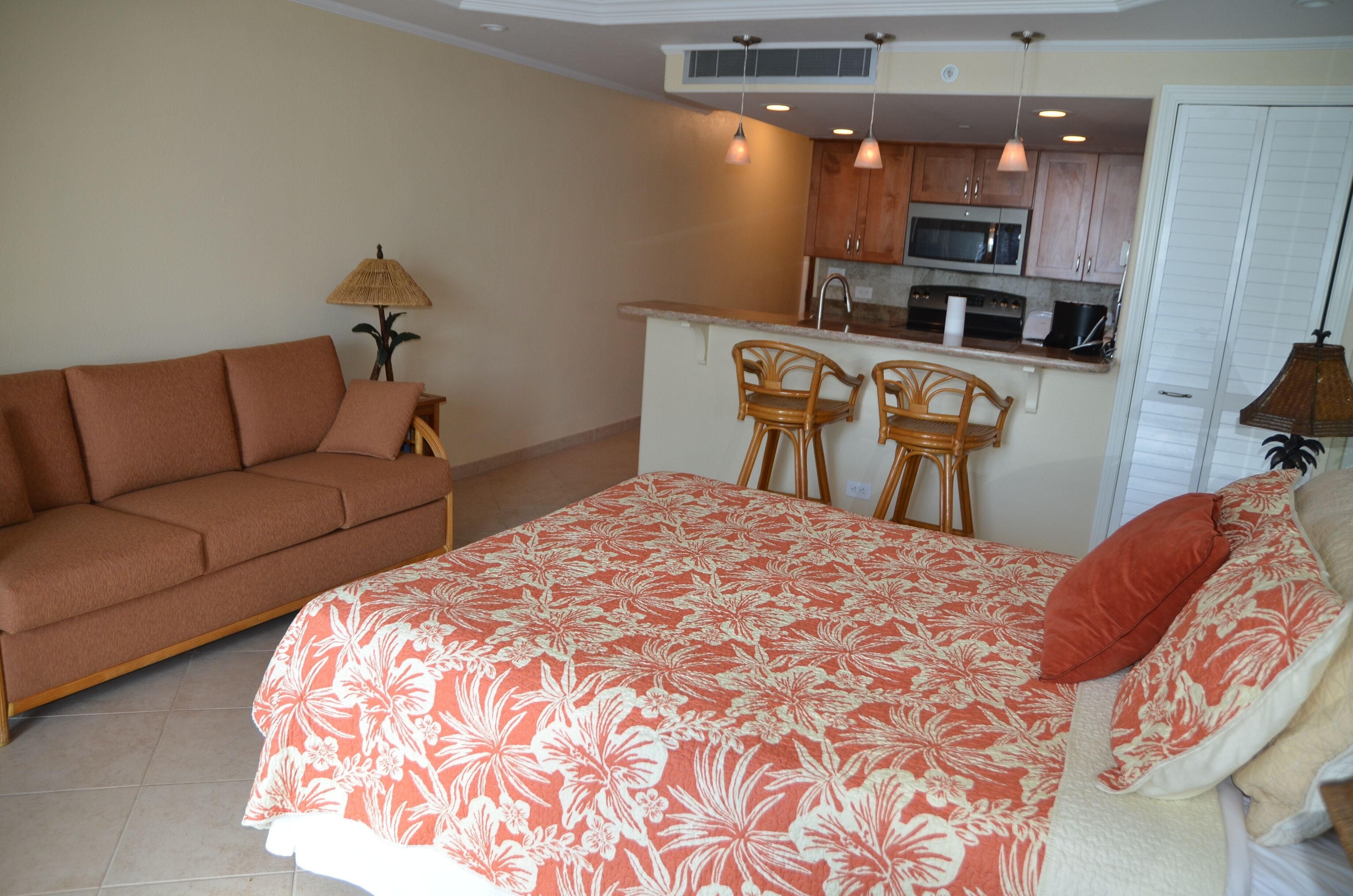 A compact living space with a sofa, a bed with a floral bedspread, two bar stools, a small kitchen area with a microwave, and a lamp.