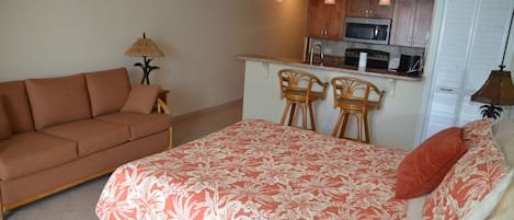 A compact living space with a sofa, a bed with a floral bedspread, two bar stools, a small kitchen area with a microwave, and a lamp.