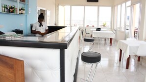 Um restaurante com área de bar, mesas e cadeiras brancas. Estantes com garrafas à esquerda.