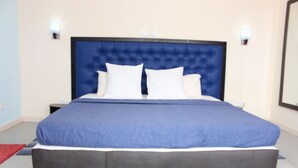 Uma cama grande com cabeceira azul, almofadas brancas e peseira cinza. Há duas mesas de cabeceira com abajures.
