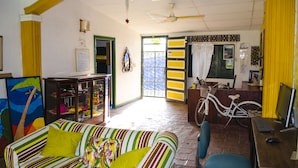 A room with a striped sofa, a bicycle, a desk with a computer, and a door leading outside. - Hostel Villa Mary (Santa Marta)