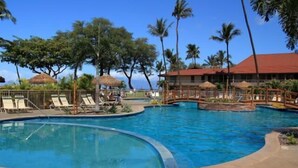 A pool area with lounge chairs and umbrellas, surrounded by palm trees and a building with a red roof. - Maui Kaanapali S #b233 1 Bedroom Condo by RedAwning (Lahaina)