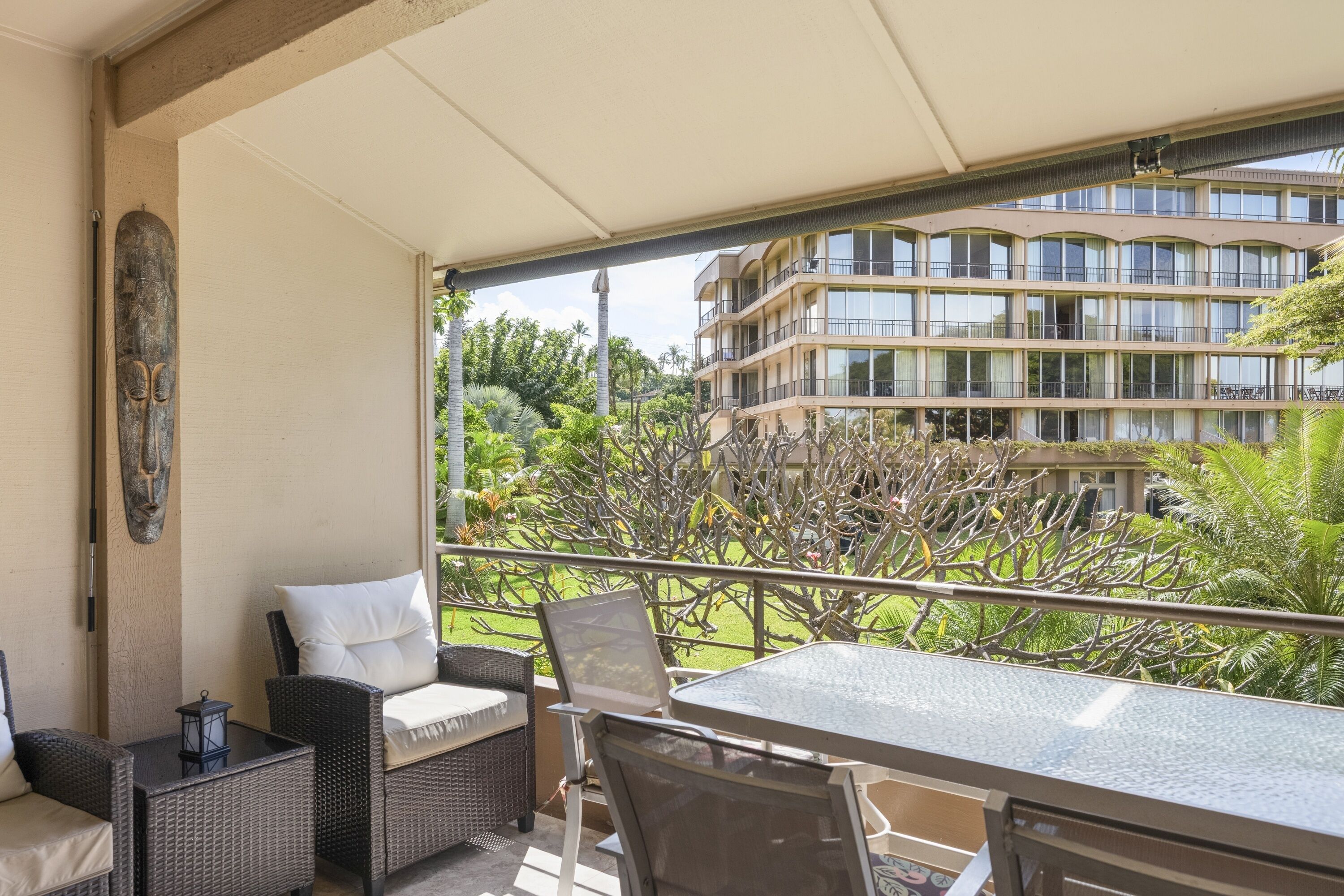 A balcony with wicker furniture, a glass table, and a view of a multi-story building and greenery.