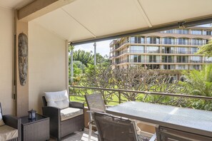 A balcony with wicker furniture, a glass table, and a view of a multi-story building and greenery. - Maui Kaanapali S #b233 1 Bedroom Condo by RedAwning (Lahaina)