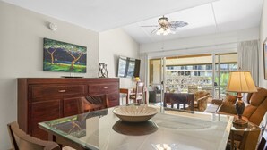 A dining area with a glass table, wooden chairs, a dark wooden cabinet, a painting on the wall, a ceiling fan, and a view through a glass door. - Maui Kaanapali S #b233 1 Bedroom Condo by RedAwning (Lahaina)