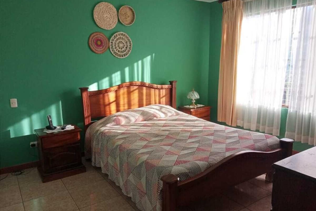 A bedroom with a wooden bed, a nightstand, a lamp, and a window with curtains.