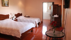 A hotel room with two beds, a small table with a vase of flowers, a TV mounted on the wall, and a bathroom visible through an open door. - Hotel Las Brumas (San Rafael)