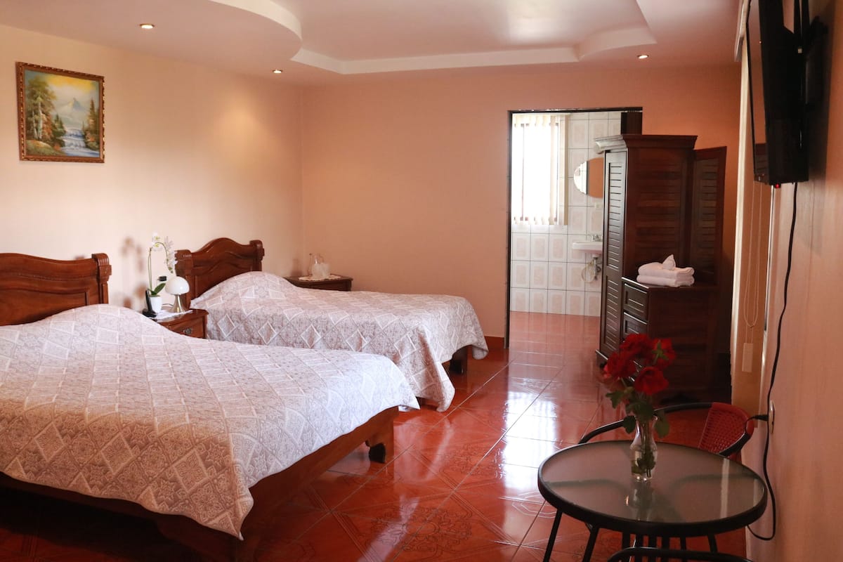 A hotel room with two beds, a small table with a vase of flowers, a TV mounted on the wall, and a bathroom visible through an open door.
