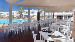 A covered outdoor dining area with white tables and chairs, overlooking a pool and a resort building. - Arena Beach (La Oliva)