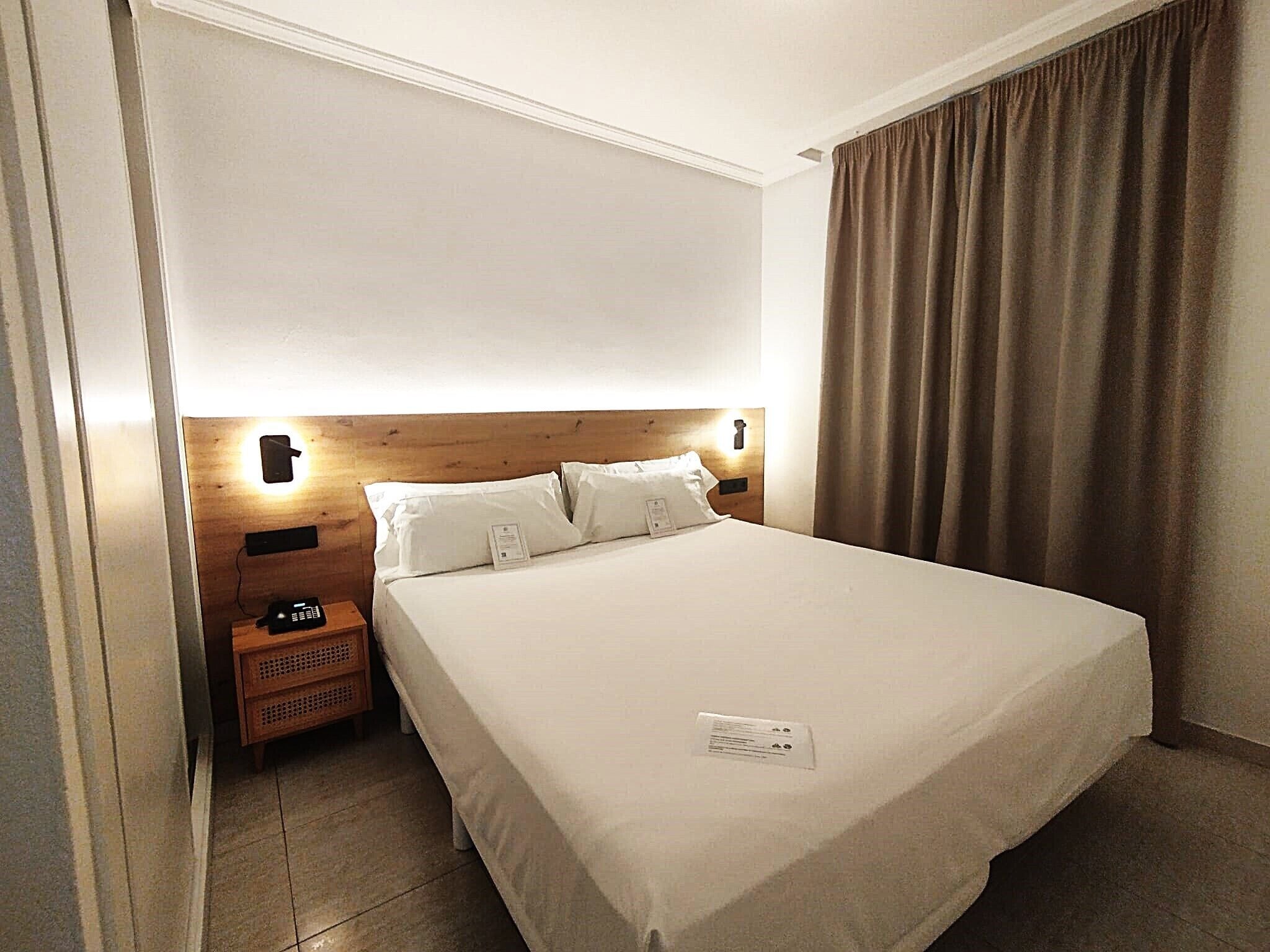 Habitación de hotel con cabecera de madera, una cama individual, mesita de noche con teléfono y una cortina en la ventana.