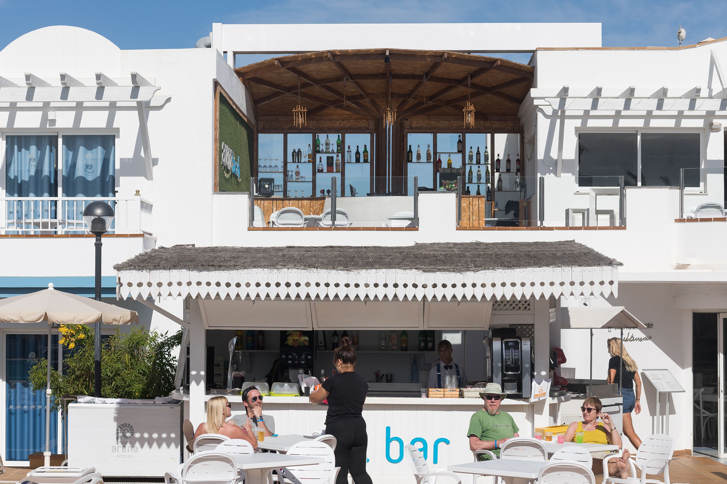Zona de bar con asientos al aire libre, una barra con un barman y vista al mar.