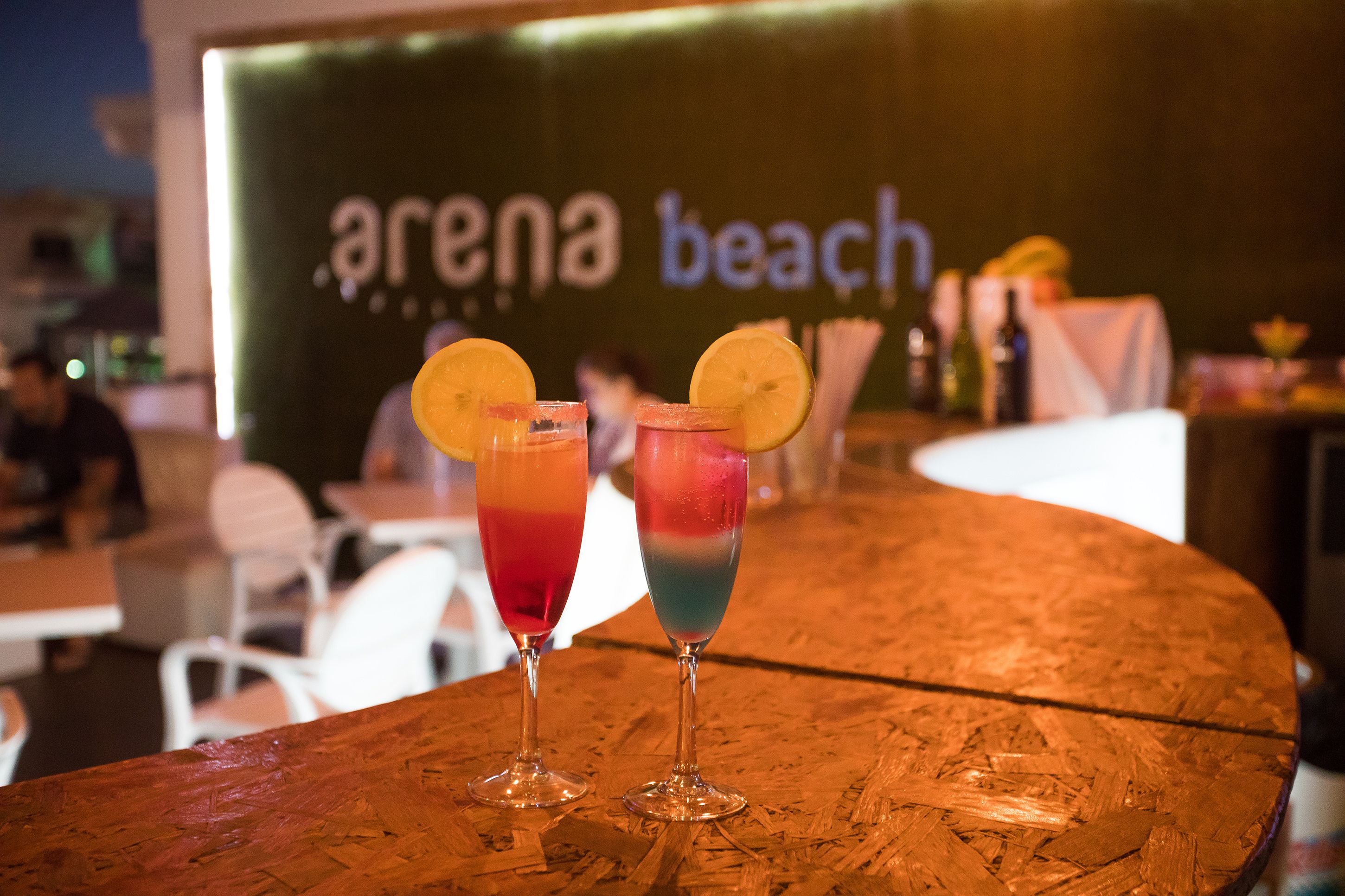 Dos cócteles coloridos con rodajas de limón sobre una barra de madera en un restaurante, con un letrero que dice 'arena beach' al fondo.