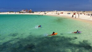 A beach with clear turquoise water, people swimming and surfing, and a building in the background. - Arena Beach (La Oliva)
