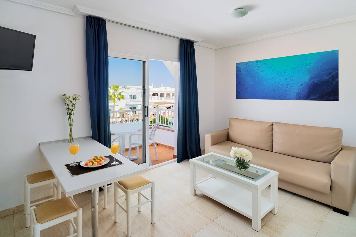 Ein modernes Wohnzimmer mit einer Couch, einem Esstisch mit Stühlen, einem Couchtisch und einem Balkon mit Blick auf Gebäude und Palmen.
