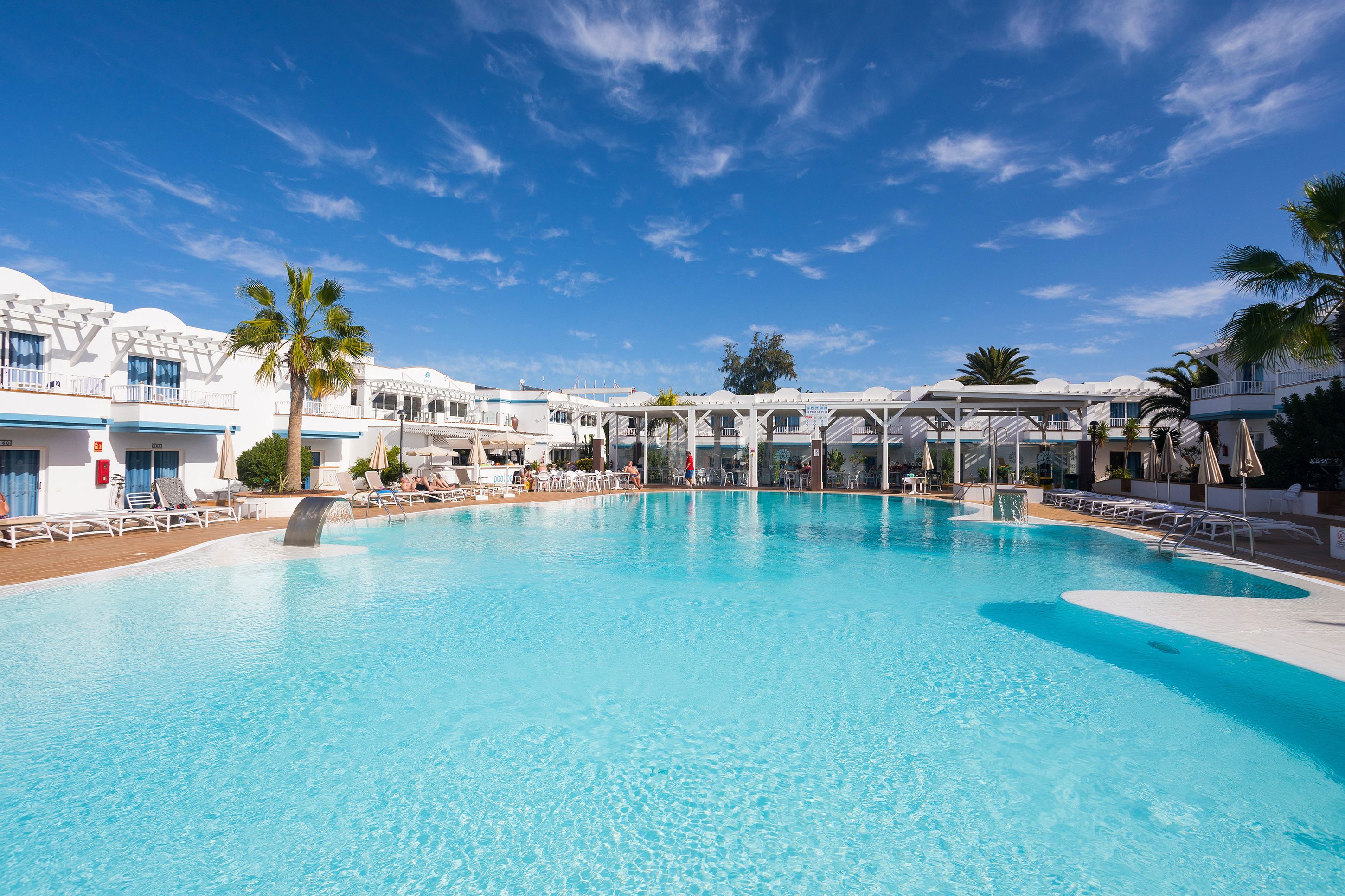 Una amplia piscina con sillones de descanso y sombrillas, rodeada de edificios blancos con detalles azules y palmeras.