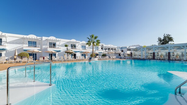 A large swimming pool with sun loungers and umbrellas, surrounded by white buildings with balconies and a clear blue sky. - Arena Beach (La Oliva)