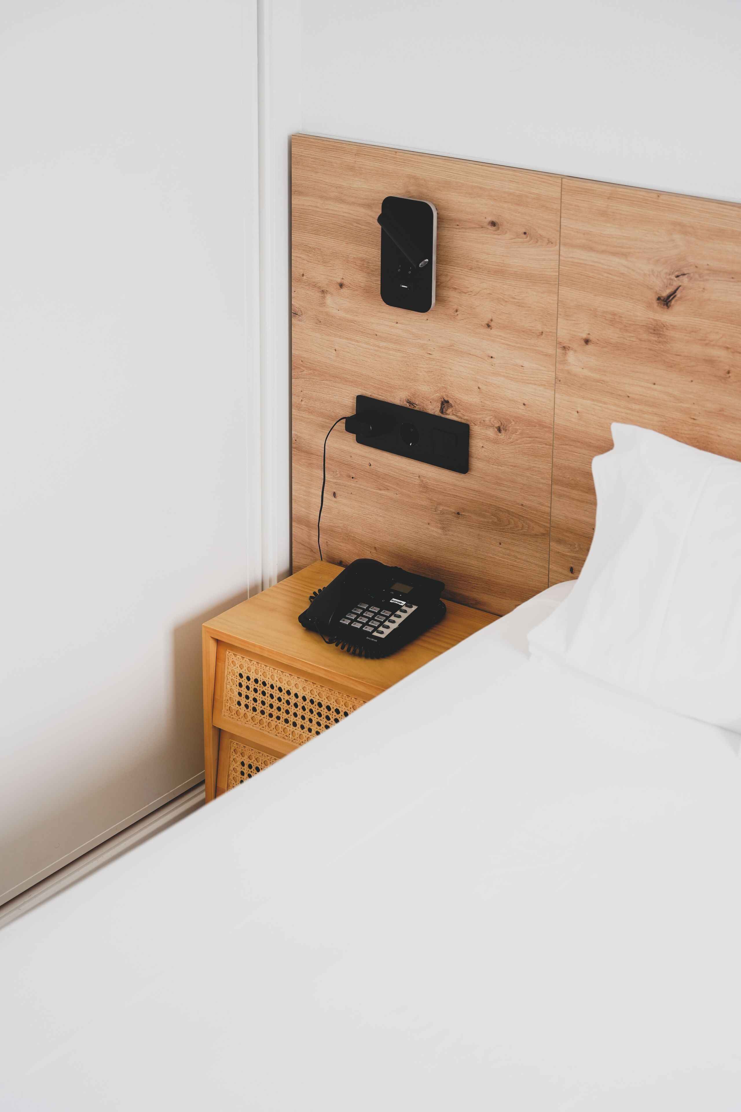 A neatly made bed with white bedding, a wooden headboard with a phone and a small shelf, and a wooden nightstand with a black object on it.