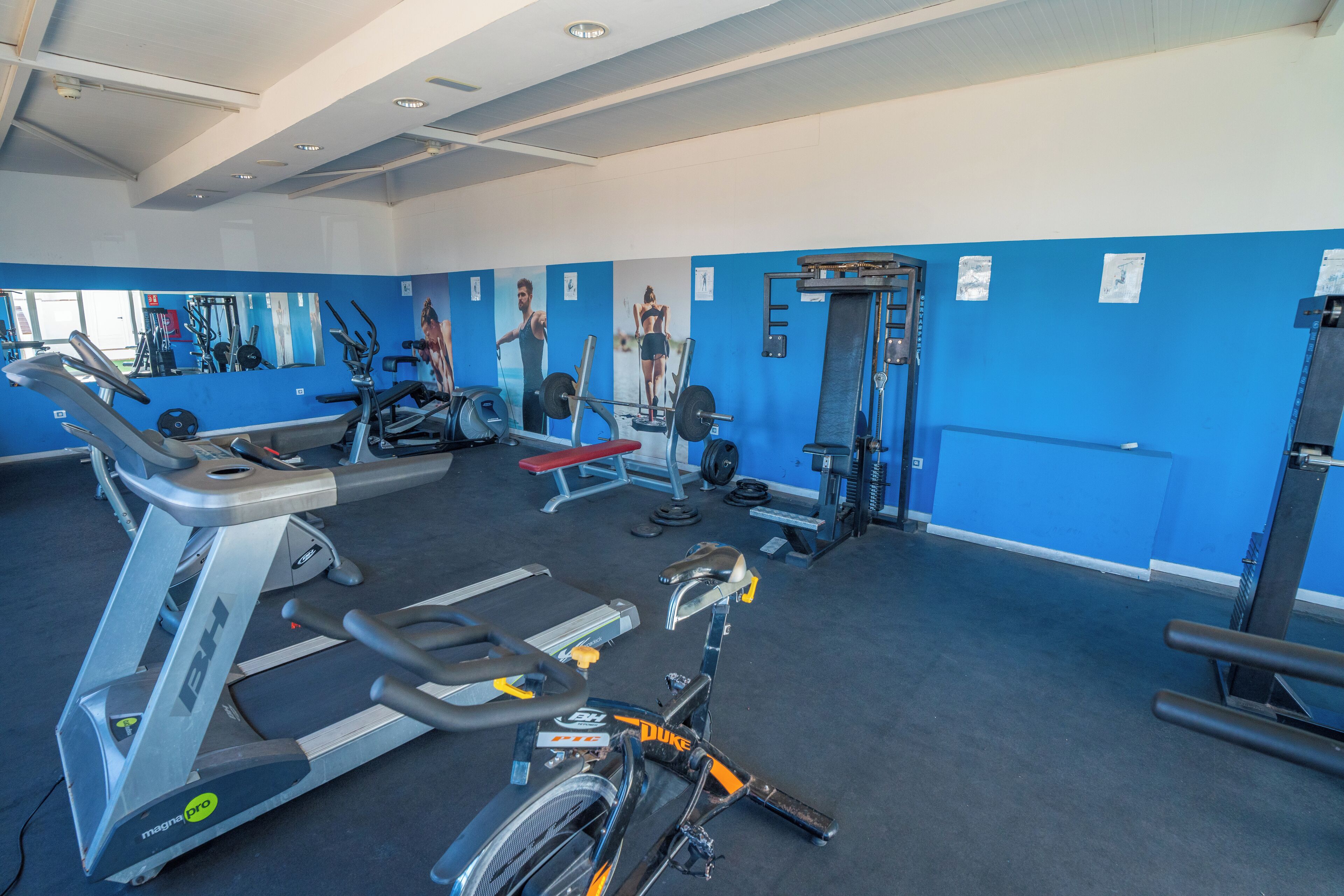 Een fitnessruimte met trainingsapparatuur, een spiegelwand en een blauwe muur met posters.