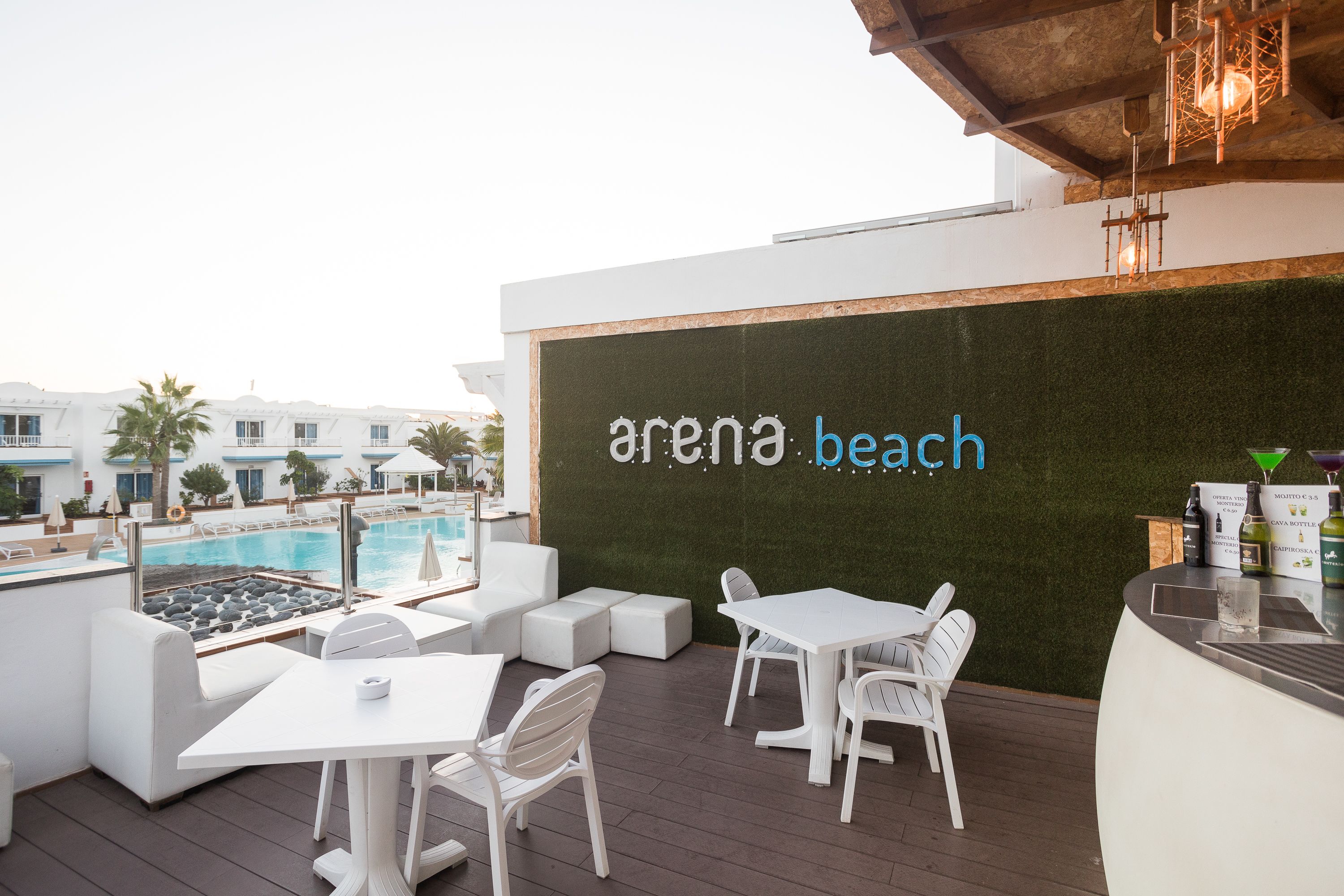 An outdoor seating area with white furniture, a pool, and a wall with 'arena beach' signage.