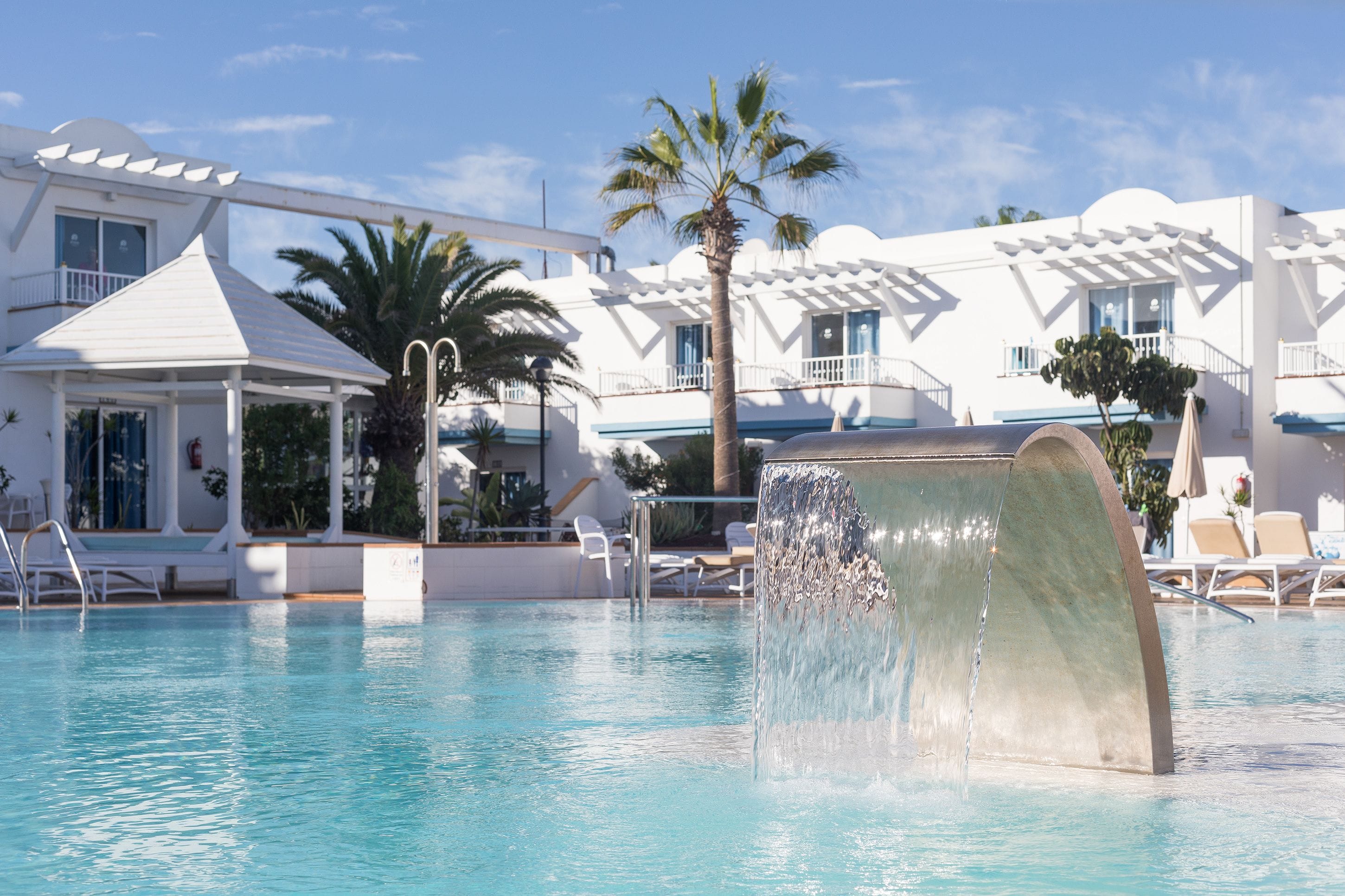 A pool with a water feature, lounge chairs, and a white building with balconies.