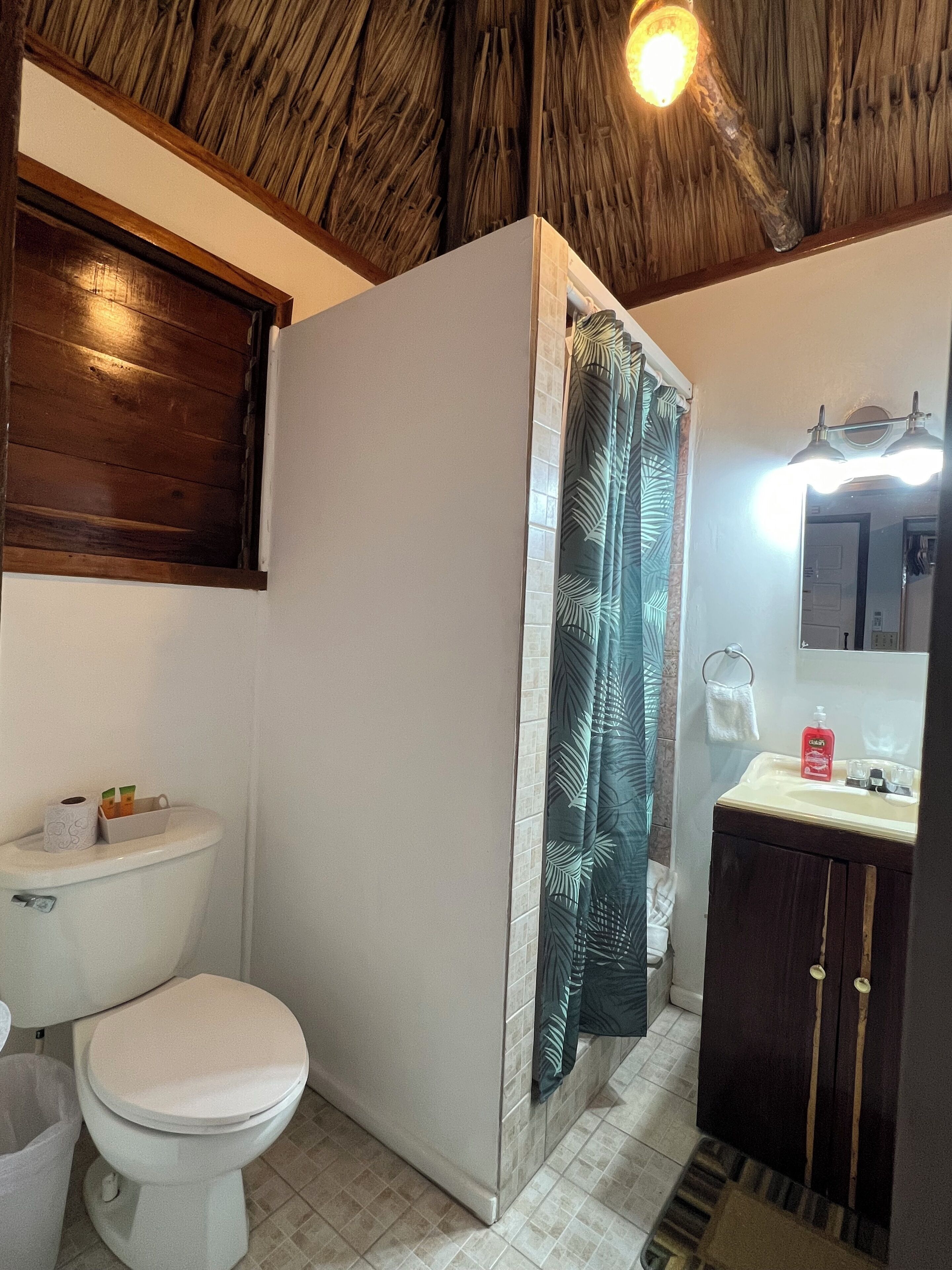 Un baño con inodoro, lavabo, ducha con cortina estampada con hojas y techo de madera.