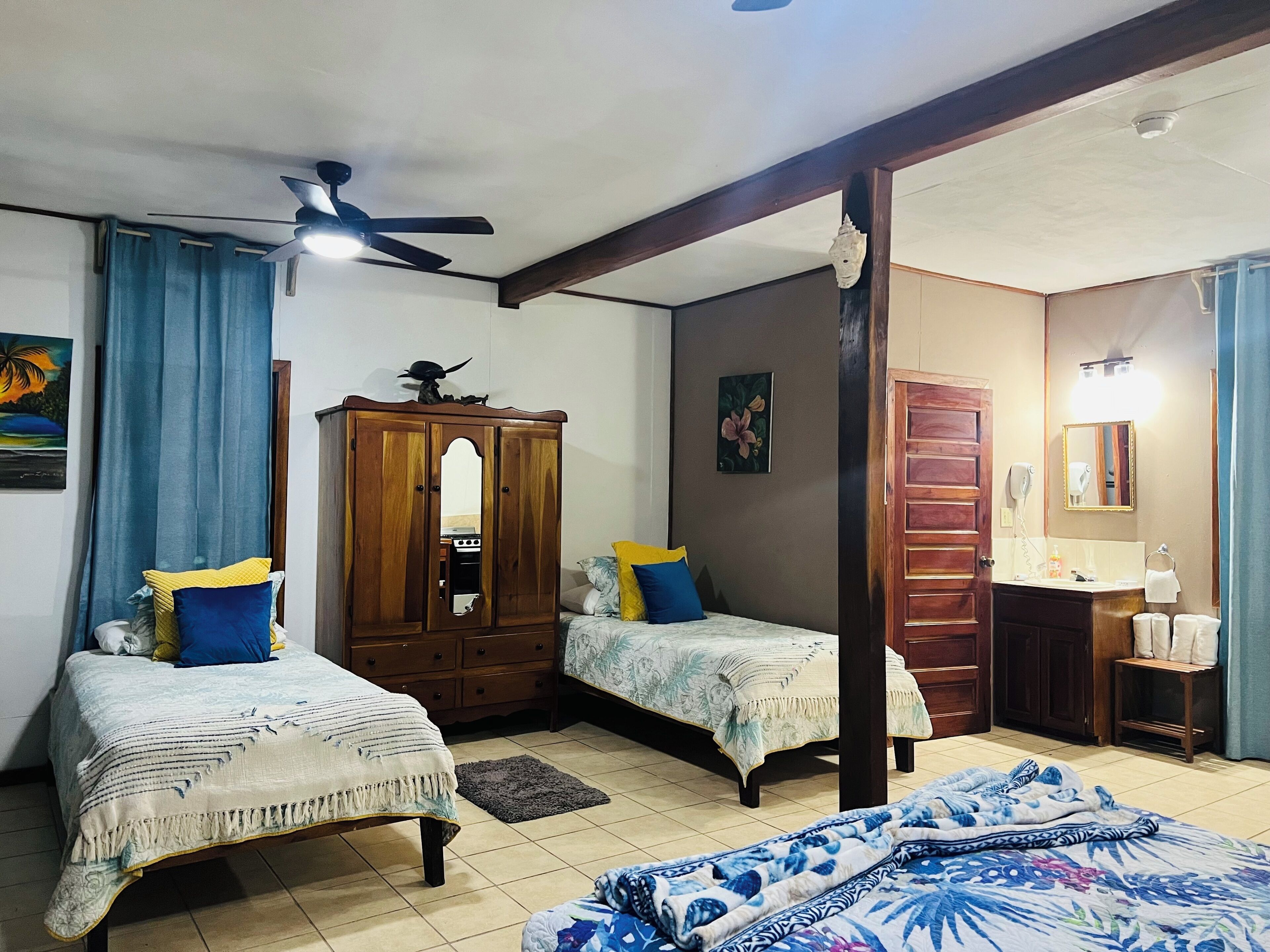 Une chambre avec deux lits, une armoire, un ventilateur de plafond et une salle de bain avec un miroir et un lavabo.