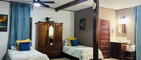 Une chambre avec deux lits, une armoire, un ventilateur de plafond et une salle de bain avec un miroir et un lavabo.