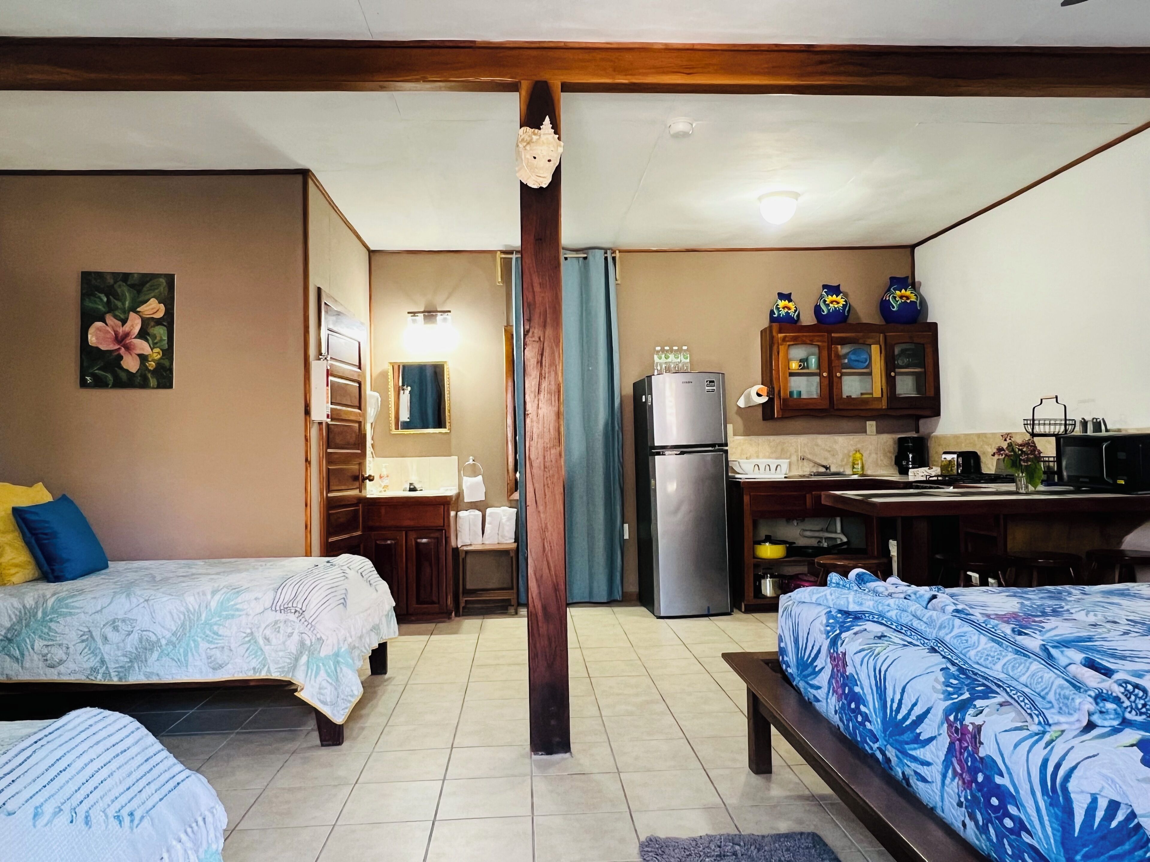 A hotel room with two beds, a kitchenette, a bathroom, and a wooden ceiling beam.