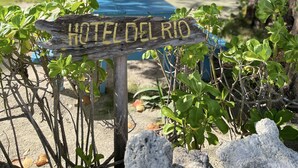 A wooden sign with 'HOTEL DEL RIO' written on it, surrounded by greenery and a pool area.