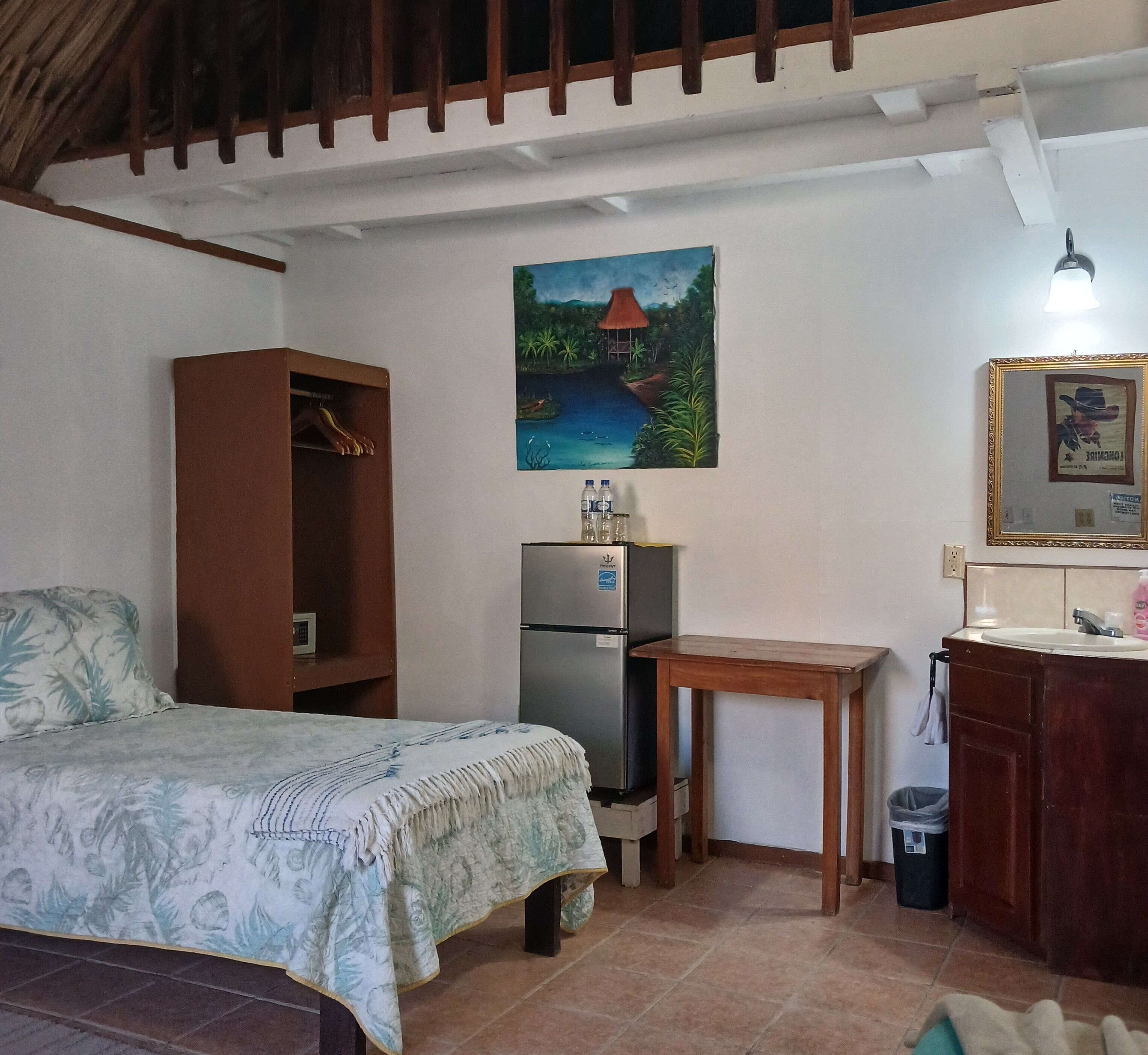 A bedroom with a bed, a wooden cabinet, a refrigerator, a small table, and a painting on the wall.