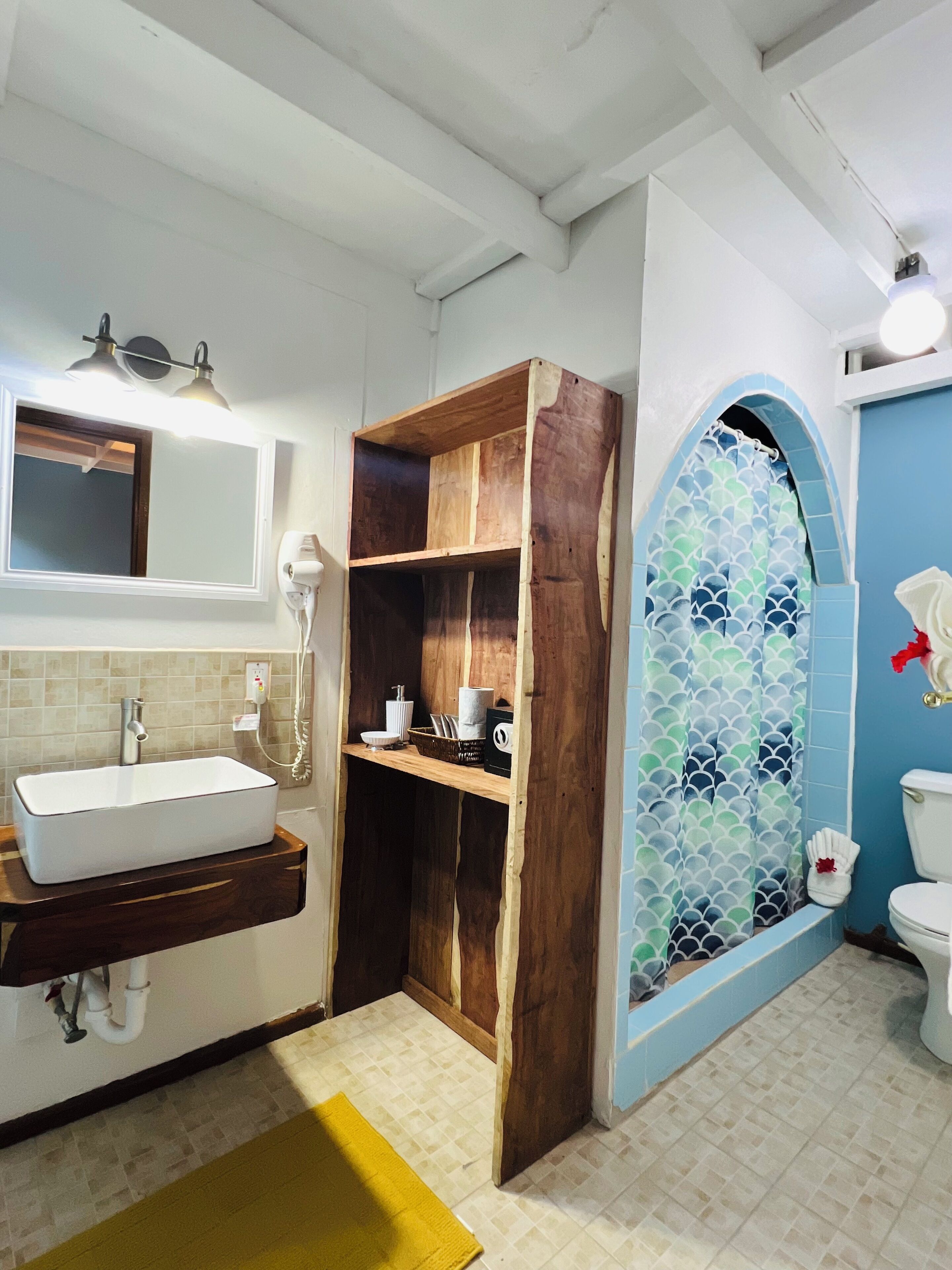 Une salle de bain avec un lavabo blanc, des étagères en bois, un miroir et une cabine de douche aux carreaux bleus.