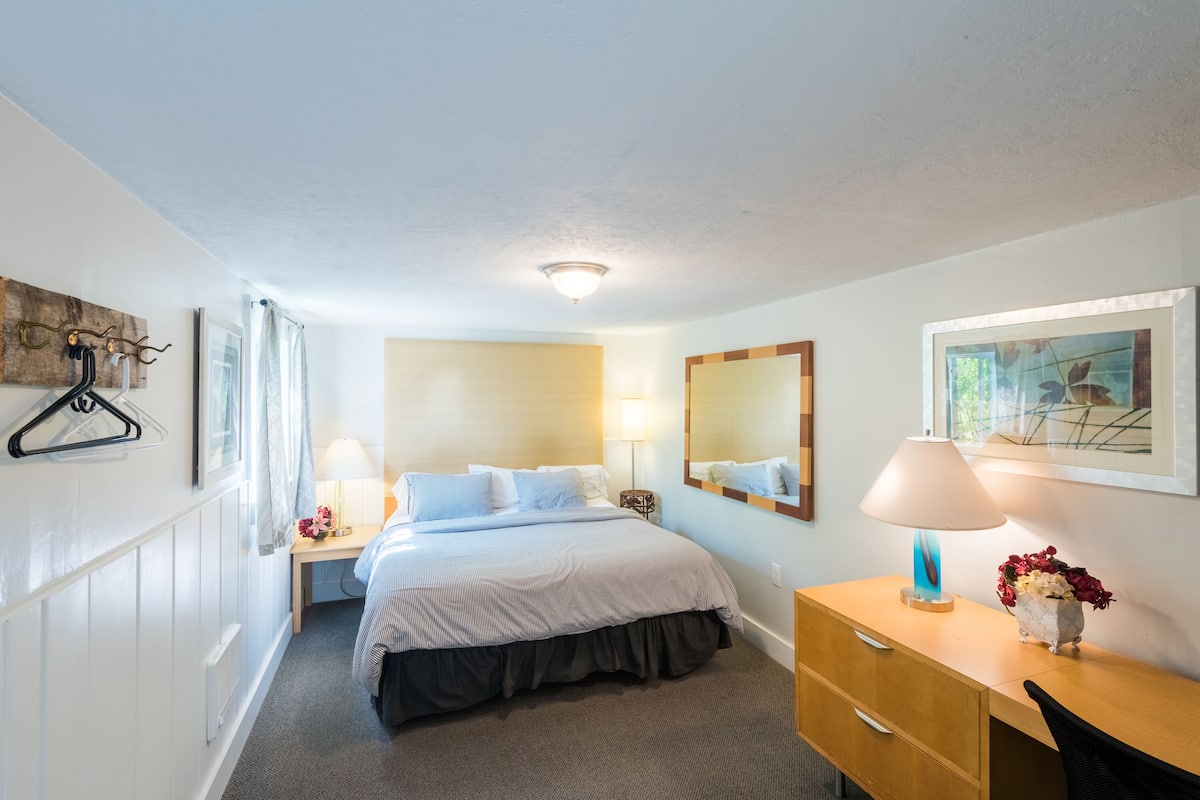 Un cuarto de hotel ordenado con una cama, mesita de noche, lámpara, cómoda y un espejo en la pared.