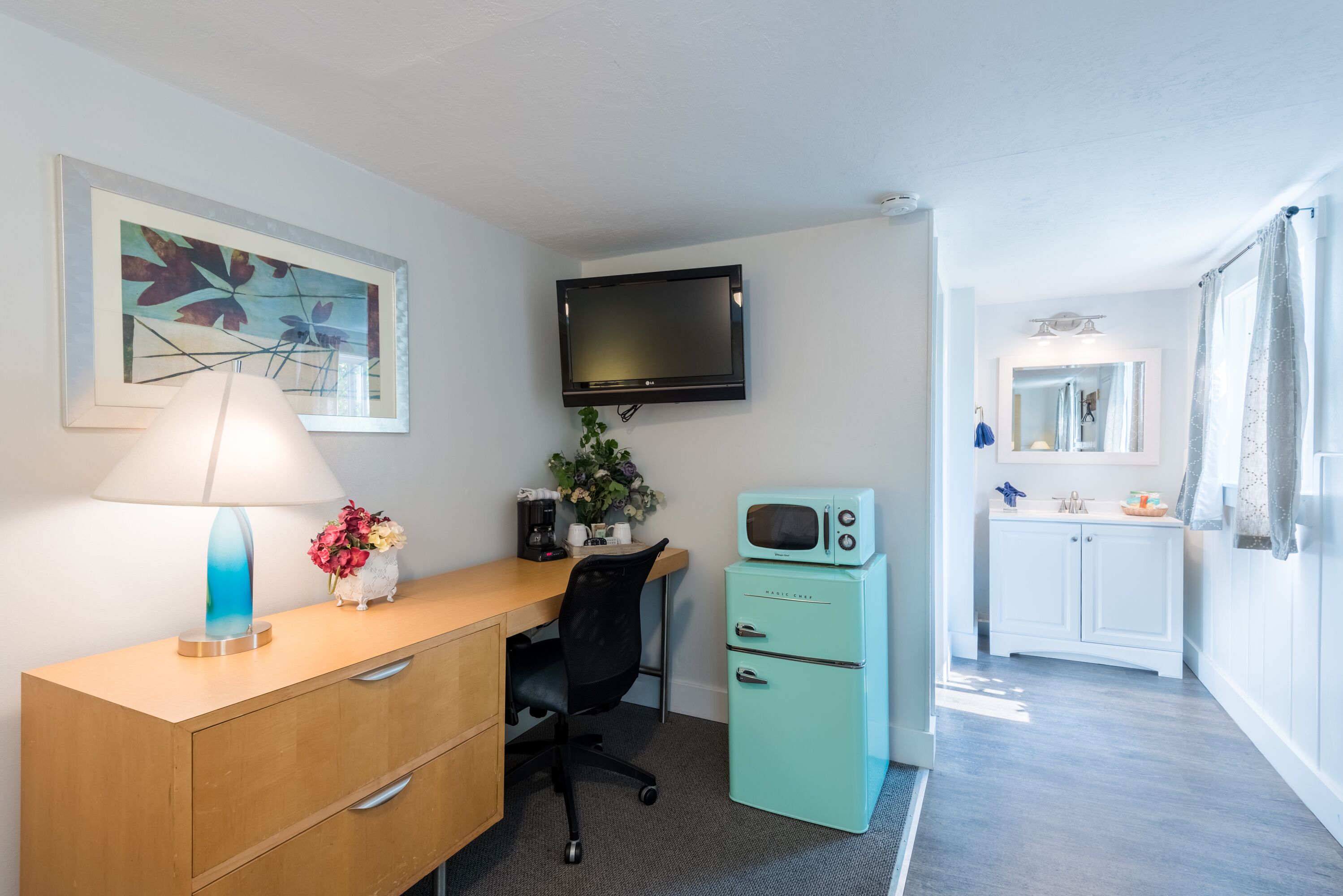 A compact hotel room with a wooden desk, a teal-colored microwave, a black office chair, a television mounted on the wall, and a small bathroom with a mirror and sink.