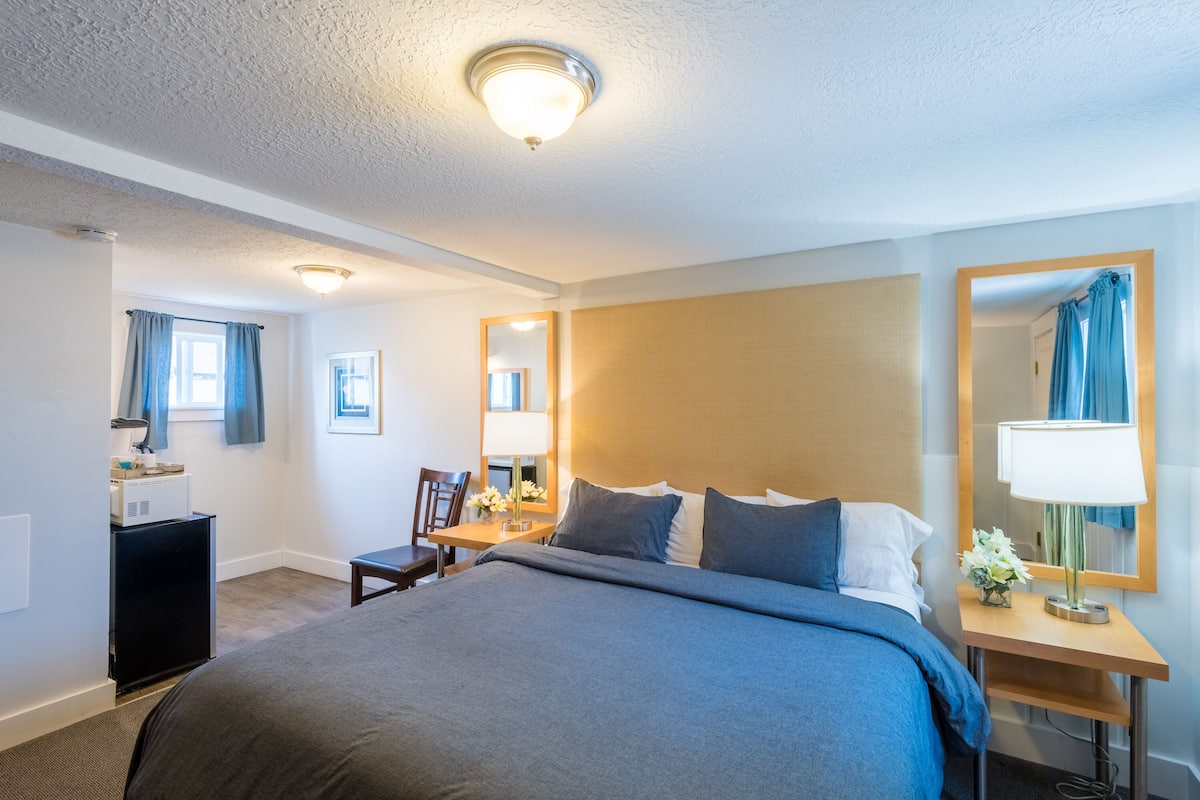 Habitación de hotel con una cama grande, un escritorio, una silla, una mesita de noche con lámpara y un espejo en la pared.